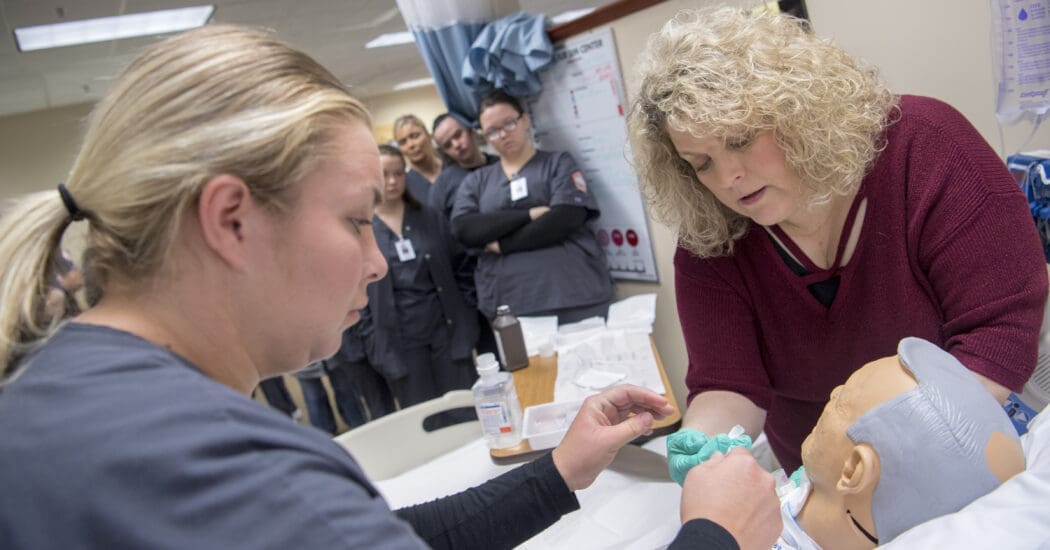 Nursing student and professor treat a patient simulator while classmates watch.