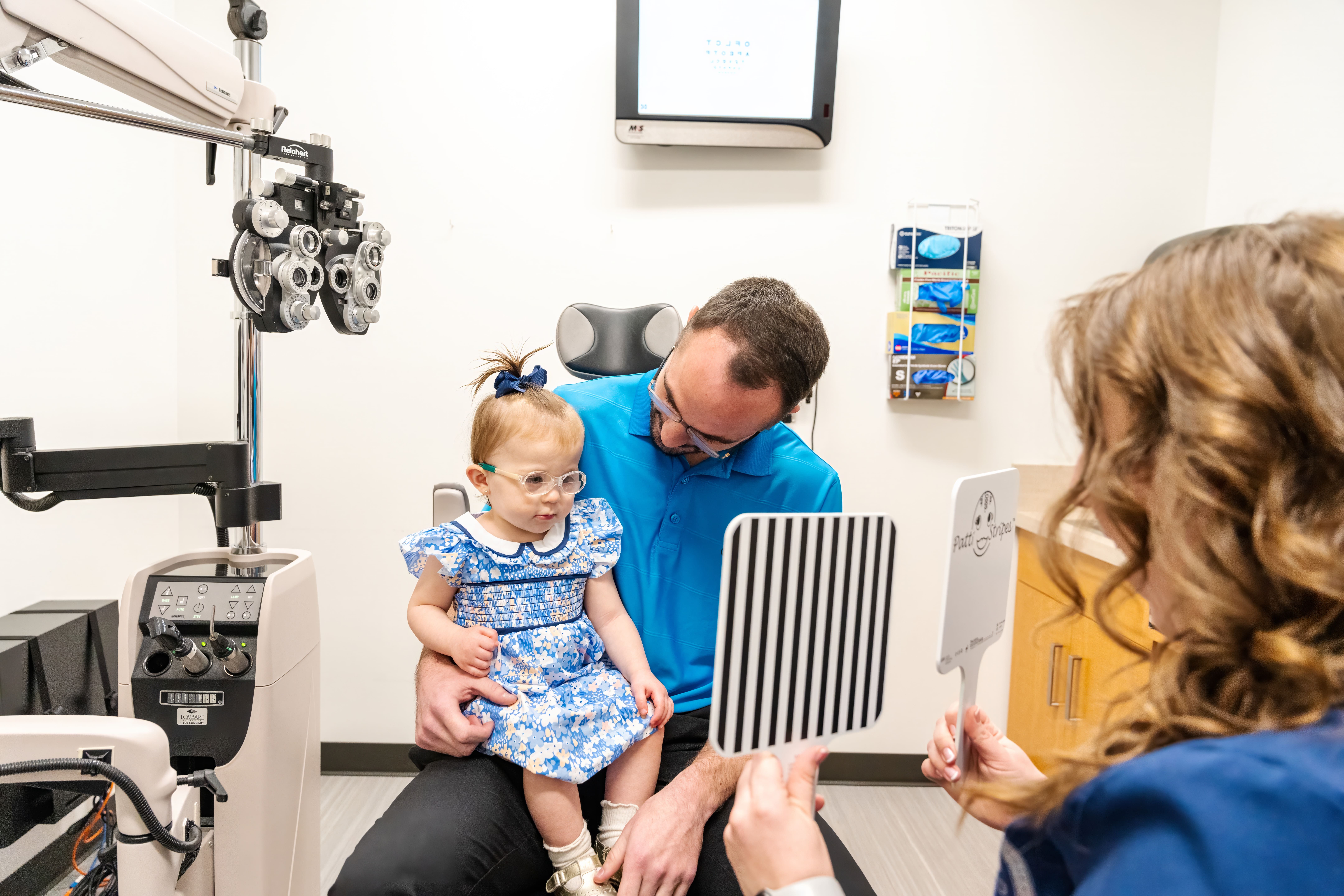 The same child sits on the man's lap in an optometry exam room. A woman in blue scrubs holds up two visual test paddles with patterns for the child to observe. The room includes vision testing equipment and educational posters.