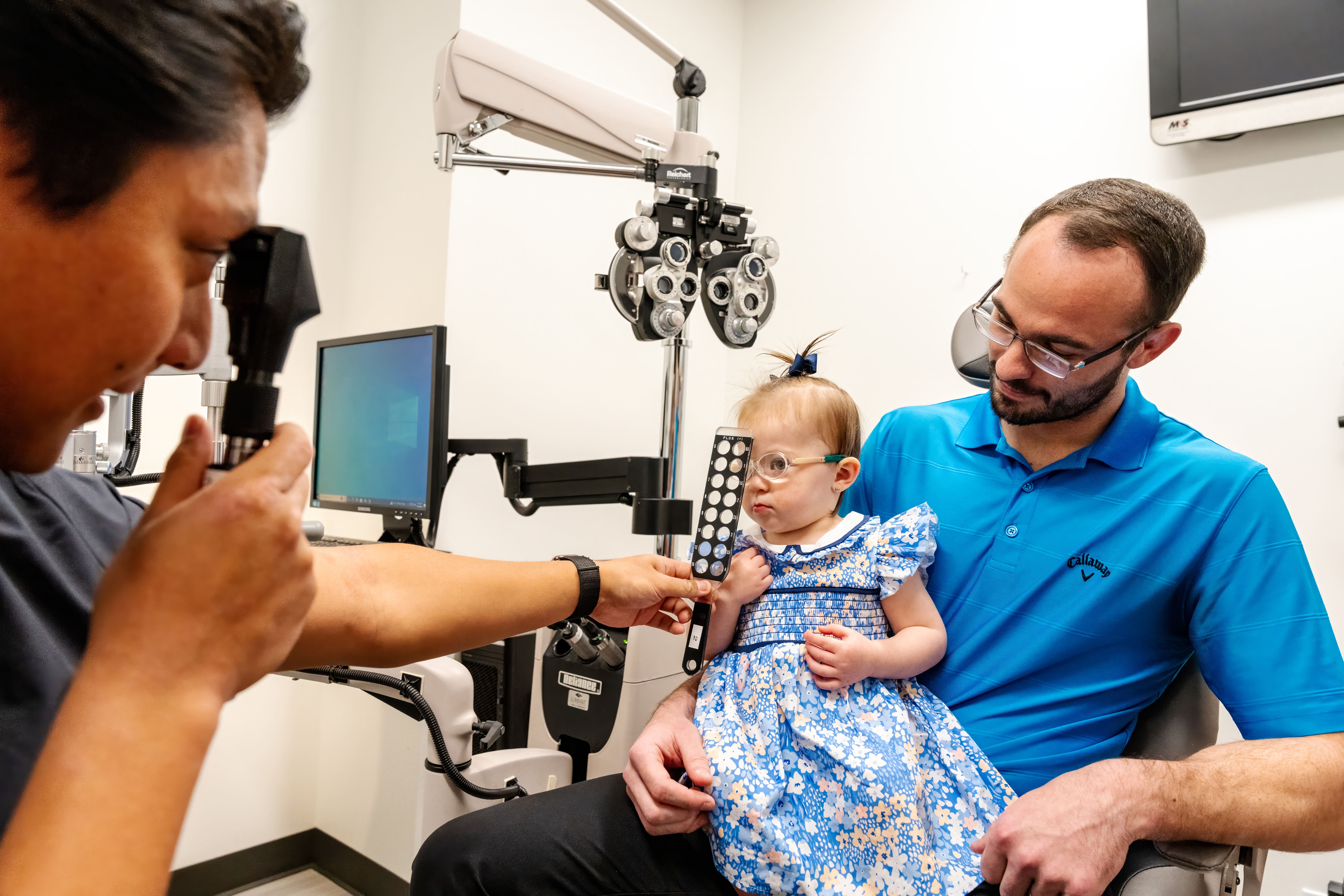 A young child wearing glasses and a blue floral dress sits on a man's lap during an eye exam. An eye care professional holds a vision testing tool in front of the child. Ophthalmology equipment is visible in the background.