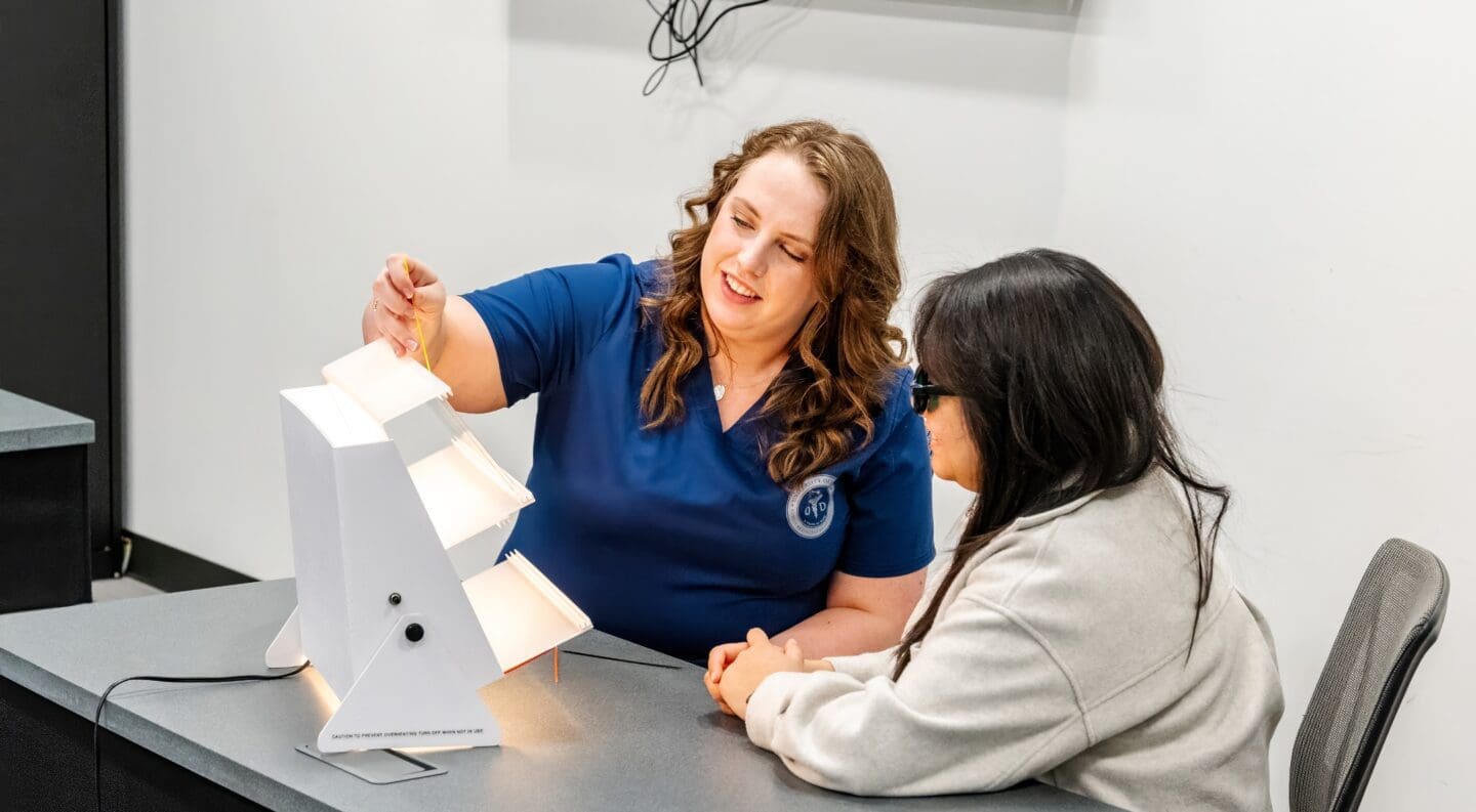 A woman in blue scrubs demonstrates a vision therapy lightboard to a seated student wearing dark glasses. They sit at a desk in a clinical classroom with a wall-mounted monitor behind them.