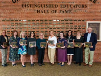 Distinguished Educators inductees lined up in front of the hall of fame wall, holding their plaques.