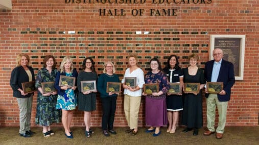 Distinguished Educators inductees lined up in front of the hall of fame wall, holding their plaques.
